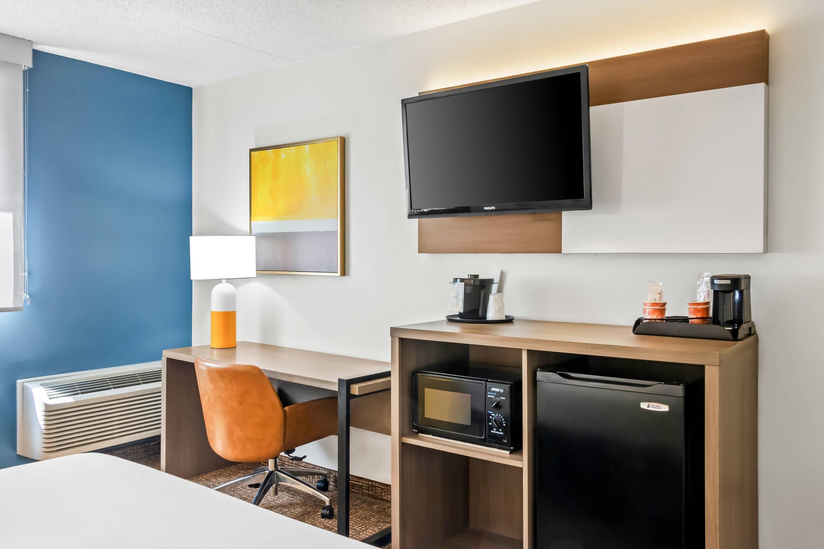Habitación de hotel con escritorio, silla, televisor de pantalla plana, microondas, refrigerador y cafetera.