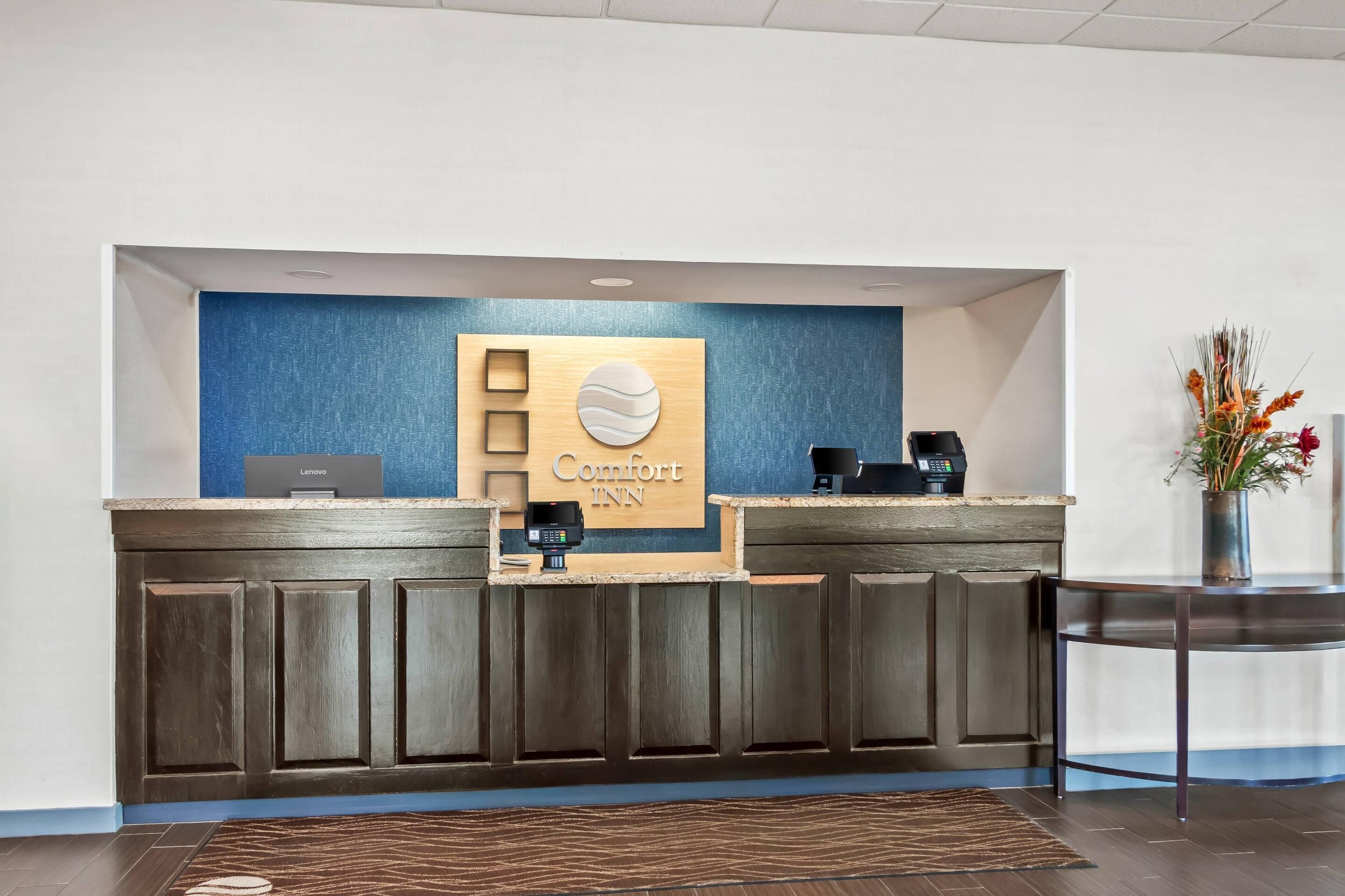 A reception desk with a blue wall, a wooden panel with 'Comfort Inn' signage, a Lenovo computer, a coffee machine, and a payment terminal.