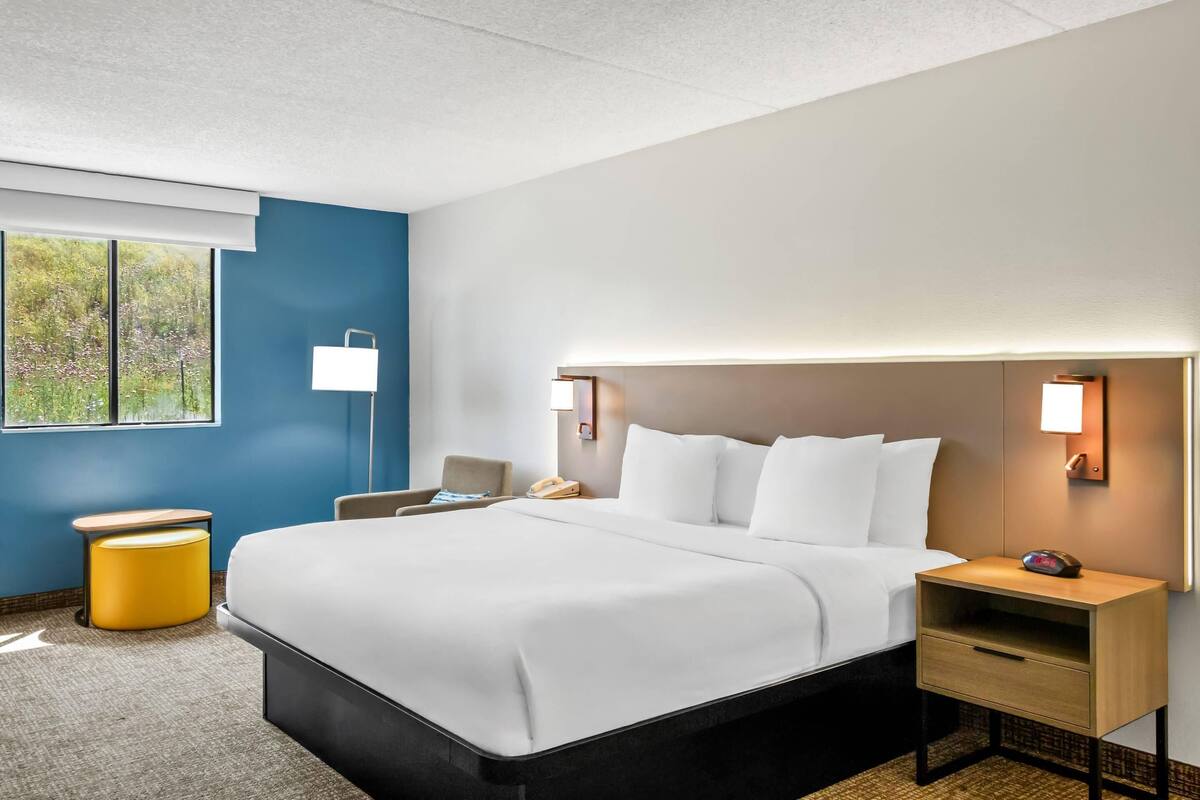 Habitación de hotel con una cama grande, mesitas de noche, una silla y ventana con vistas a zonas verdes.