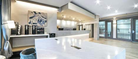 A modern hotel lobby with a marble reception desk, coffee machines, and a large abstract painting.