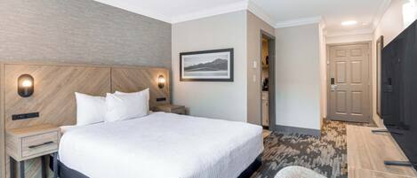 A hotel room with a large bed, a wooden headboard, a nightstand, a desk, a chair, a flat-screen TV, and a framed picture on the wall.