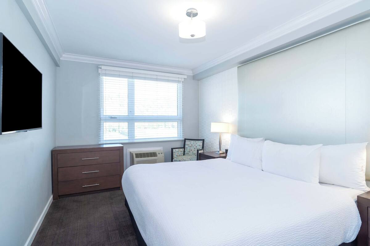 A hotel room with a large bed, a flat-screen TV mounted on the wall, a window with blinds, a bedside table with a lamp, and a small dresser.