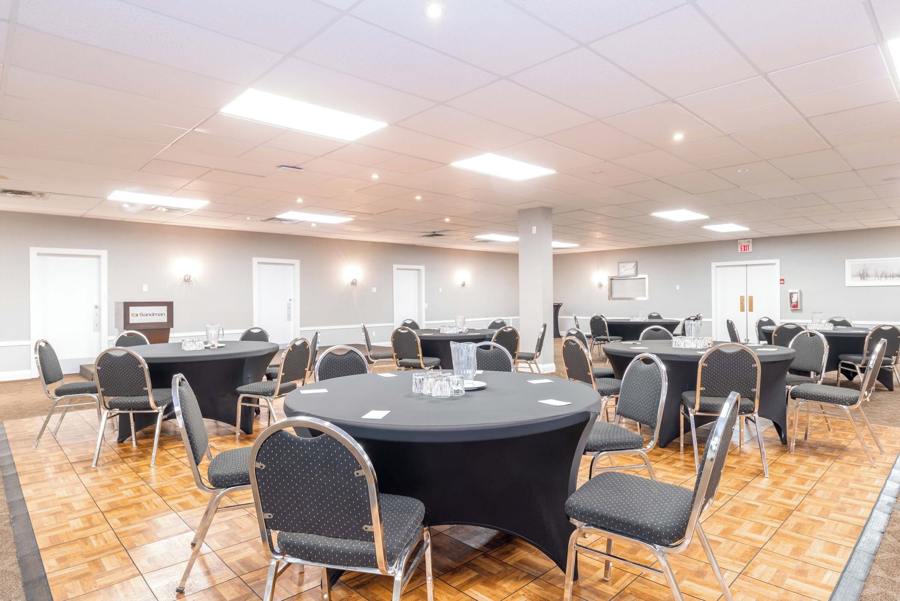 Un salón de conferencias con varias mesas redondas, cada una rodeada de sillas, en un espacio bien iluminado con suelo de baldosas.
