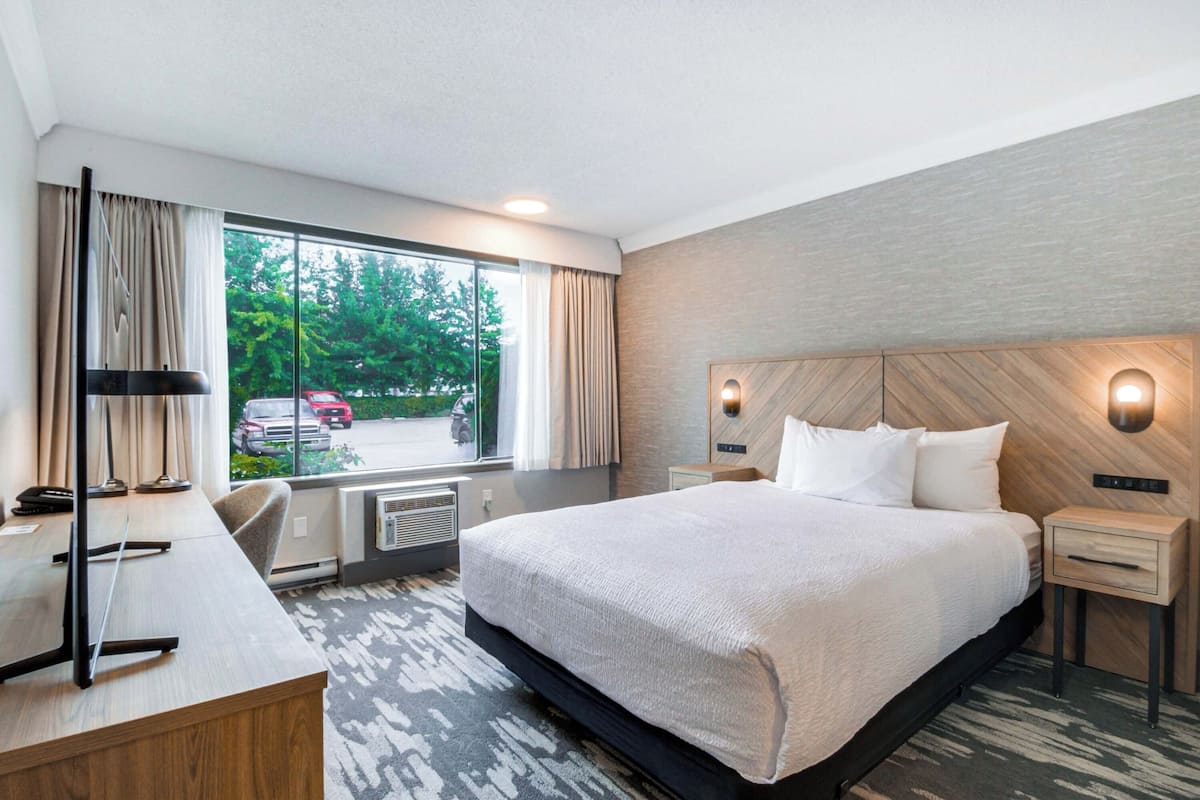 A hotel room with a large bed, a desk with a chair, a window with curtains, and a view of the outside.
