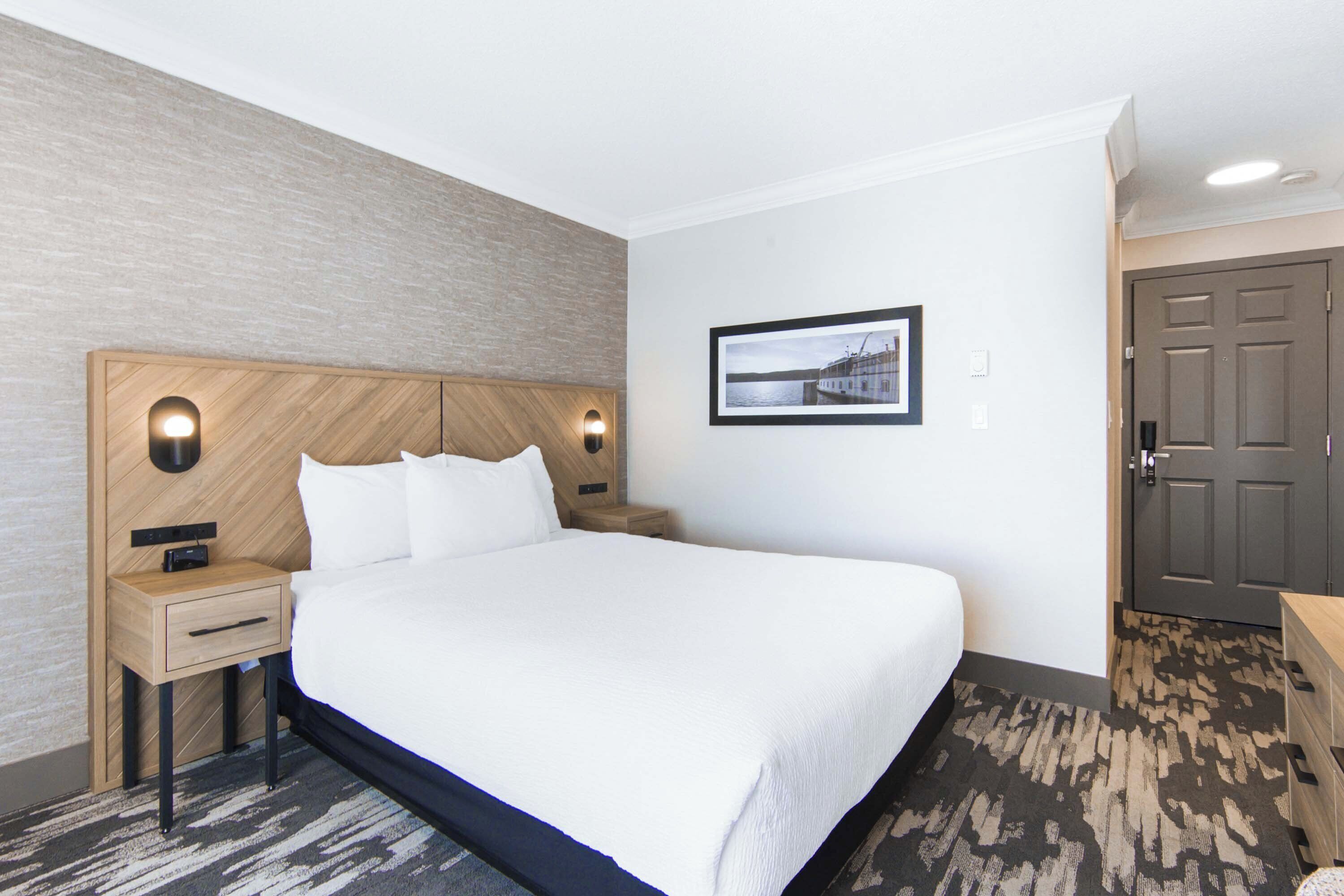 A hotel room with a large bed, a wooden headboard, a nightstand, a framed picture on the wall, and a door.