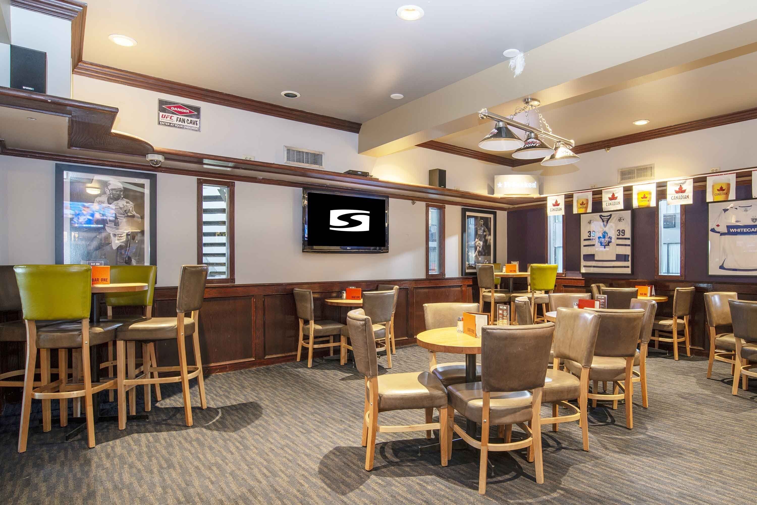 A hotel lobby with wooden chairs, tables, a mounted TV, and a sign indicating a UFC fan cave.