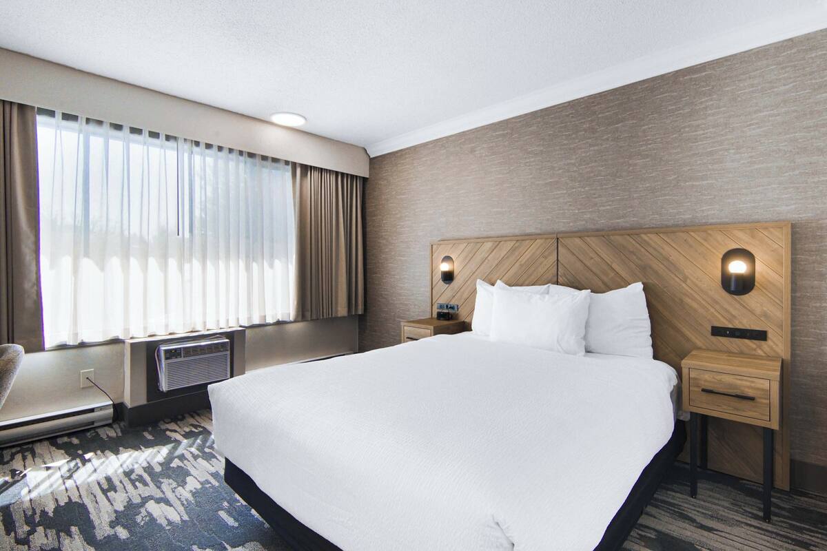 A hotel room with a large bed, a wooden headboard, two bedside tables, a chair, and a large window with curtains.