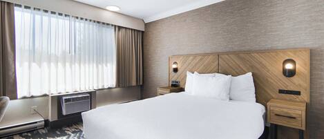A hotel room with a large bed, a wooden headboard, two bedside tables, a chair, and a large window with curtains.