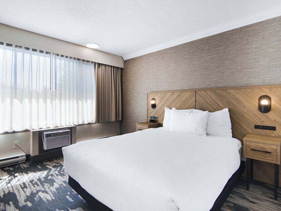 Een hotelkamer met een groot bed, een houten hoofdbord, twee nachtkastjes, een stoel en een groot raam met gordijnen.