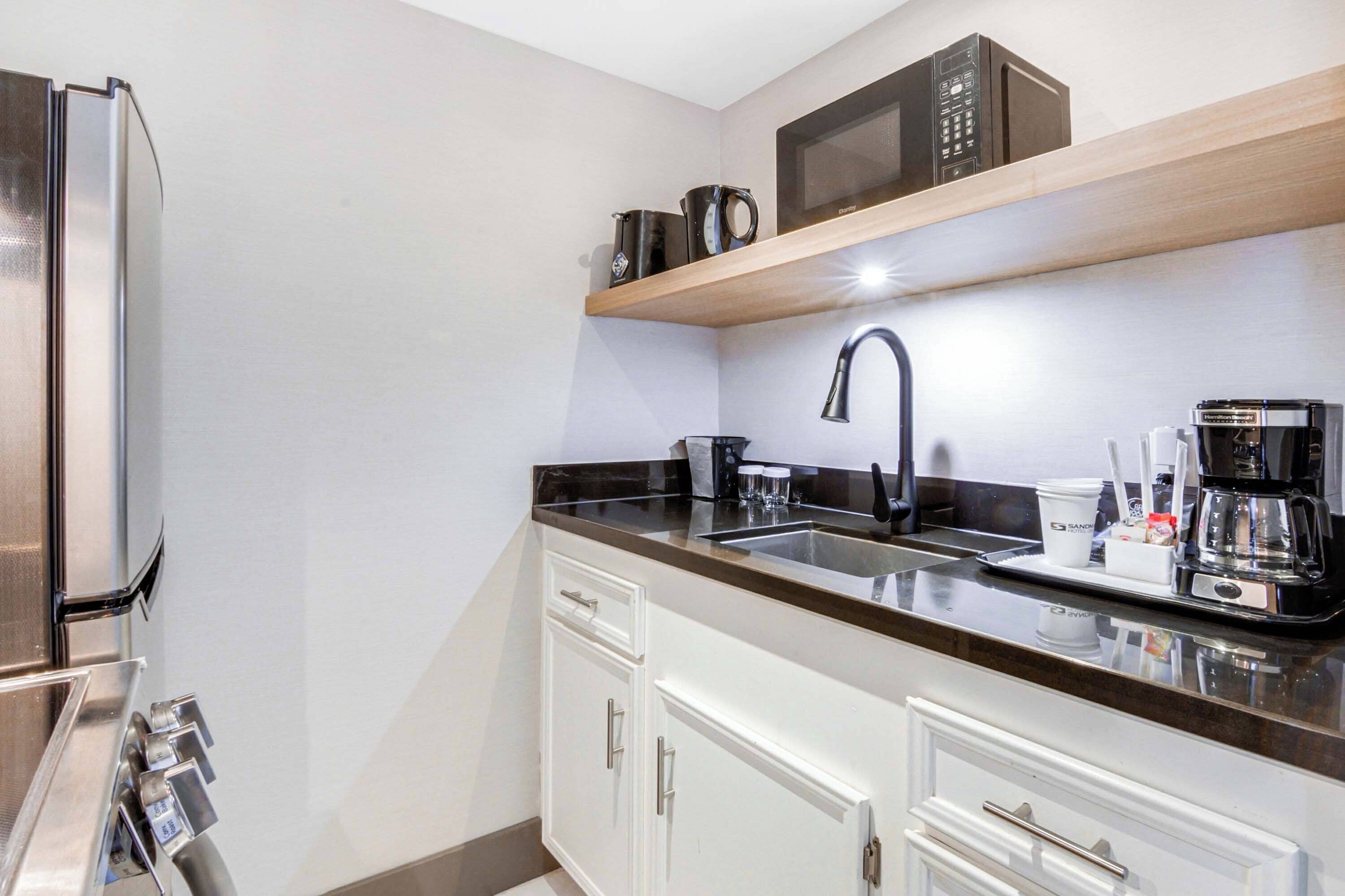A compact kitchen with a stainless steel refrigerator, a microwave on a wooden shelf, a coffee maker, and a sink.