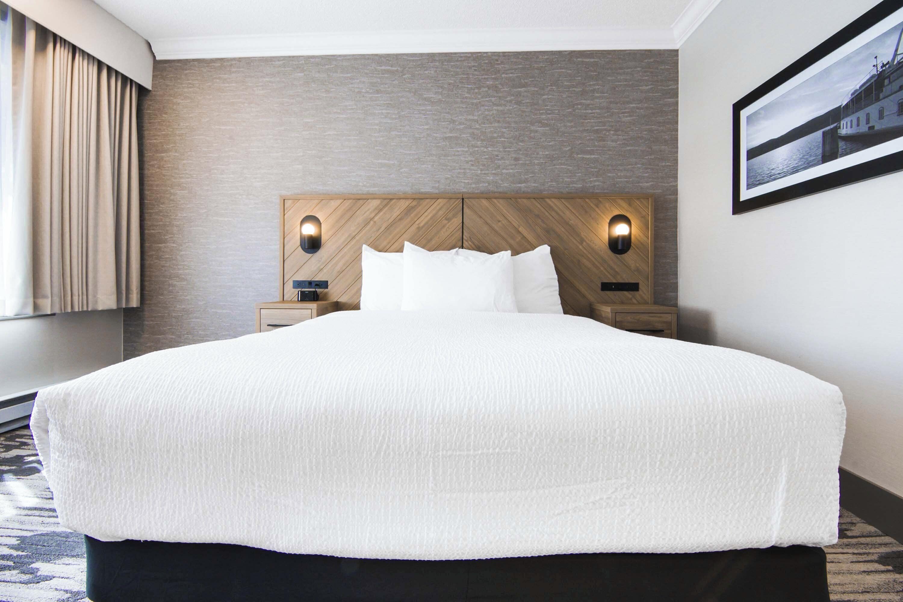 A hotel room with a large bed, wooden headboard, two bedside tables with lamps, a large window with curtains, and a framed picture on the wall.