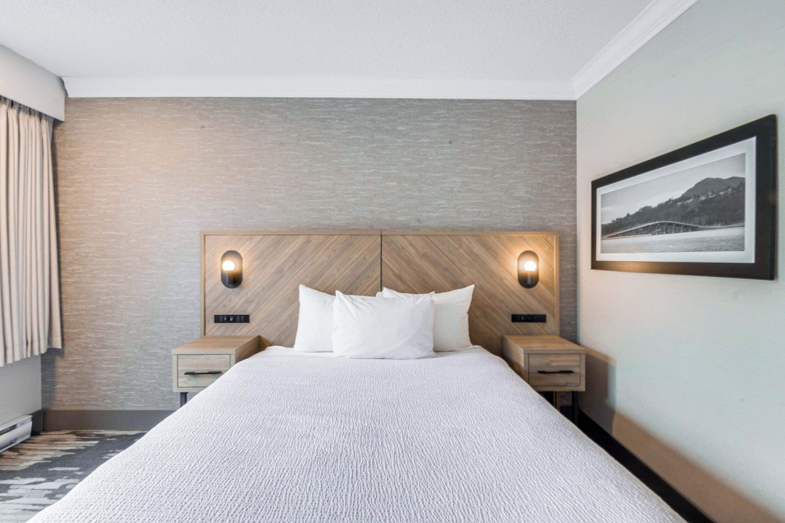 A hotel room with a large bed, two bedside tables, a headboard with integrated lighting, a framed picture on the wall, and a window with curtains.
