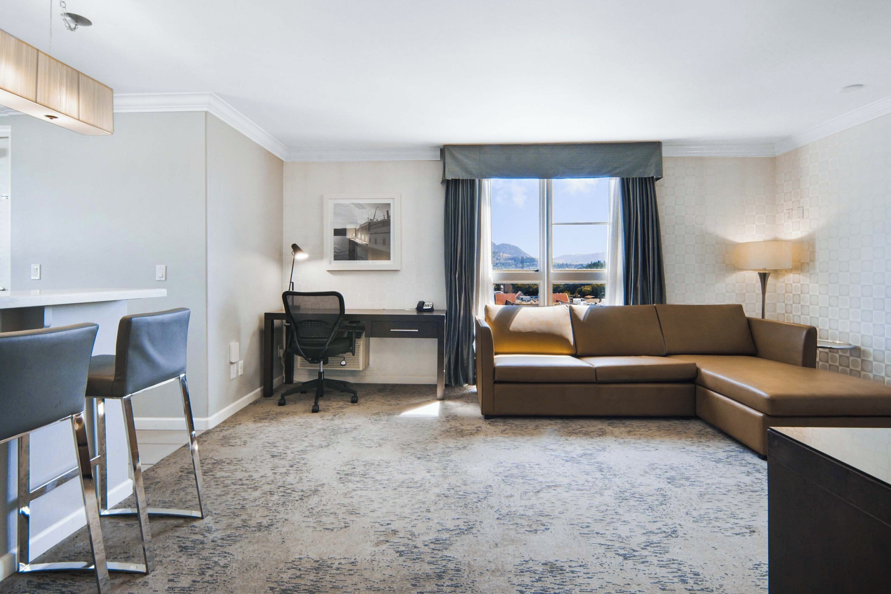 A hotel room with a desk, two chairs, a sofa, a lamp, a window with a view, and a framed picture on the wall.