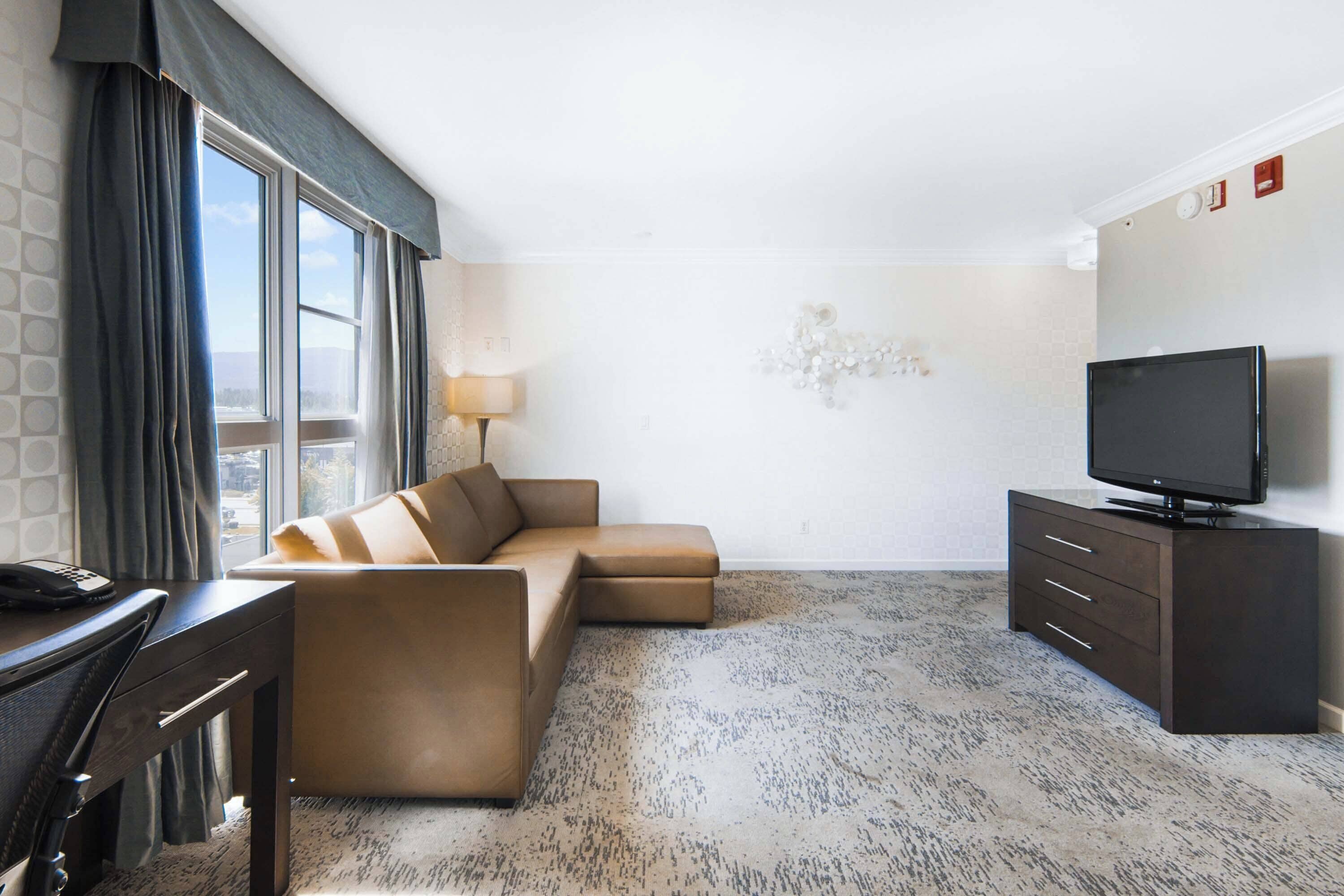 Una habitación de hotel moderna con un sofá seccional, un televisor de pantalla plana, un escritorio con teléfono y un amplio ventanal con cortinas.