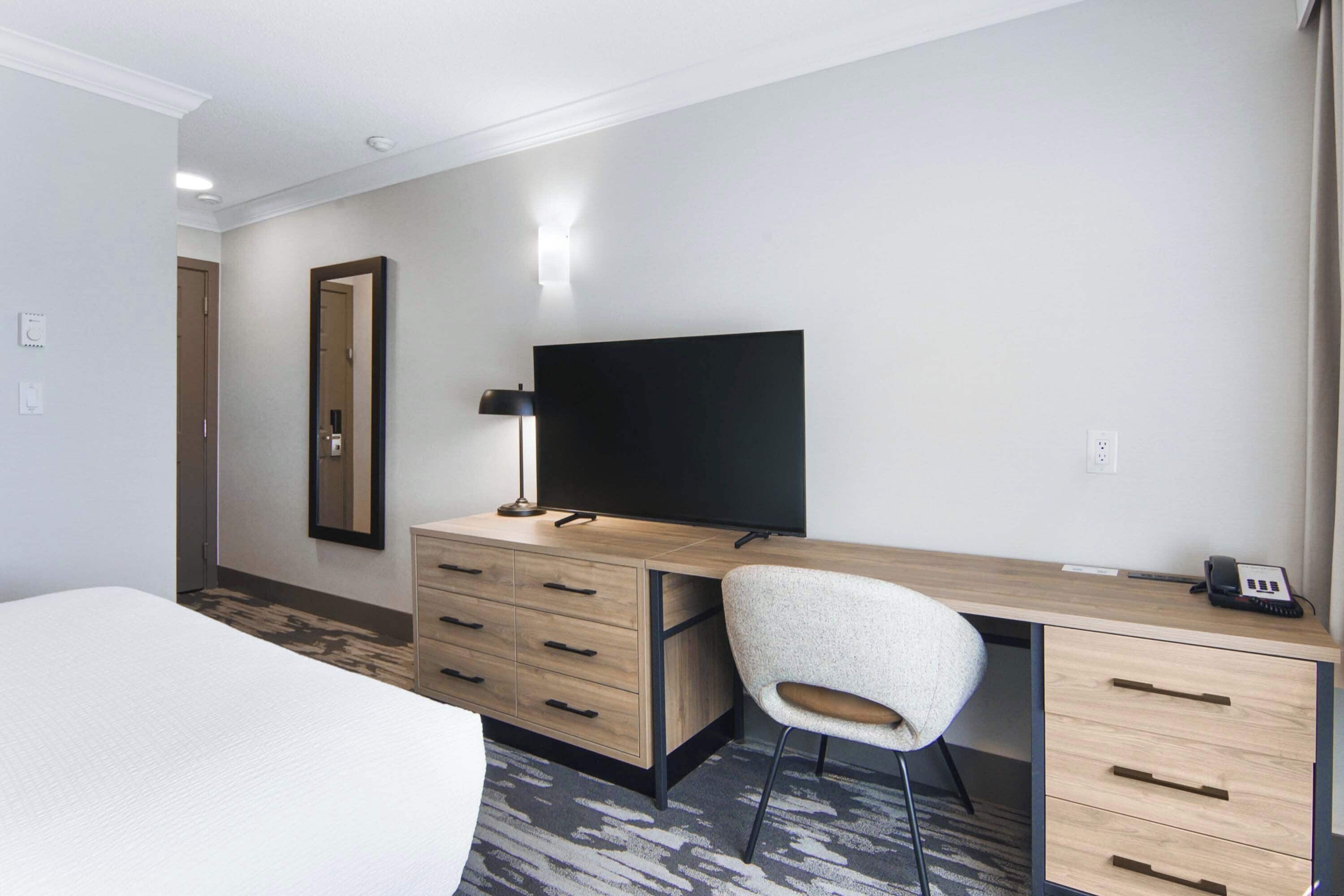 A hotel room with a bed, a desk with a computer and a lamp, a chair, and a large mirror.