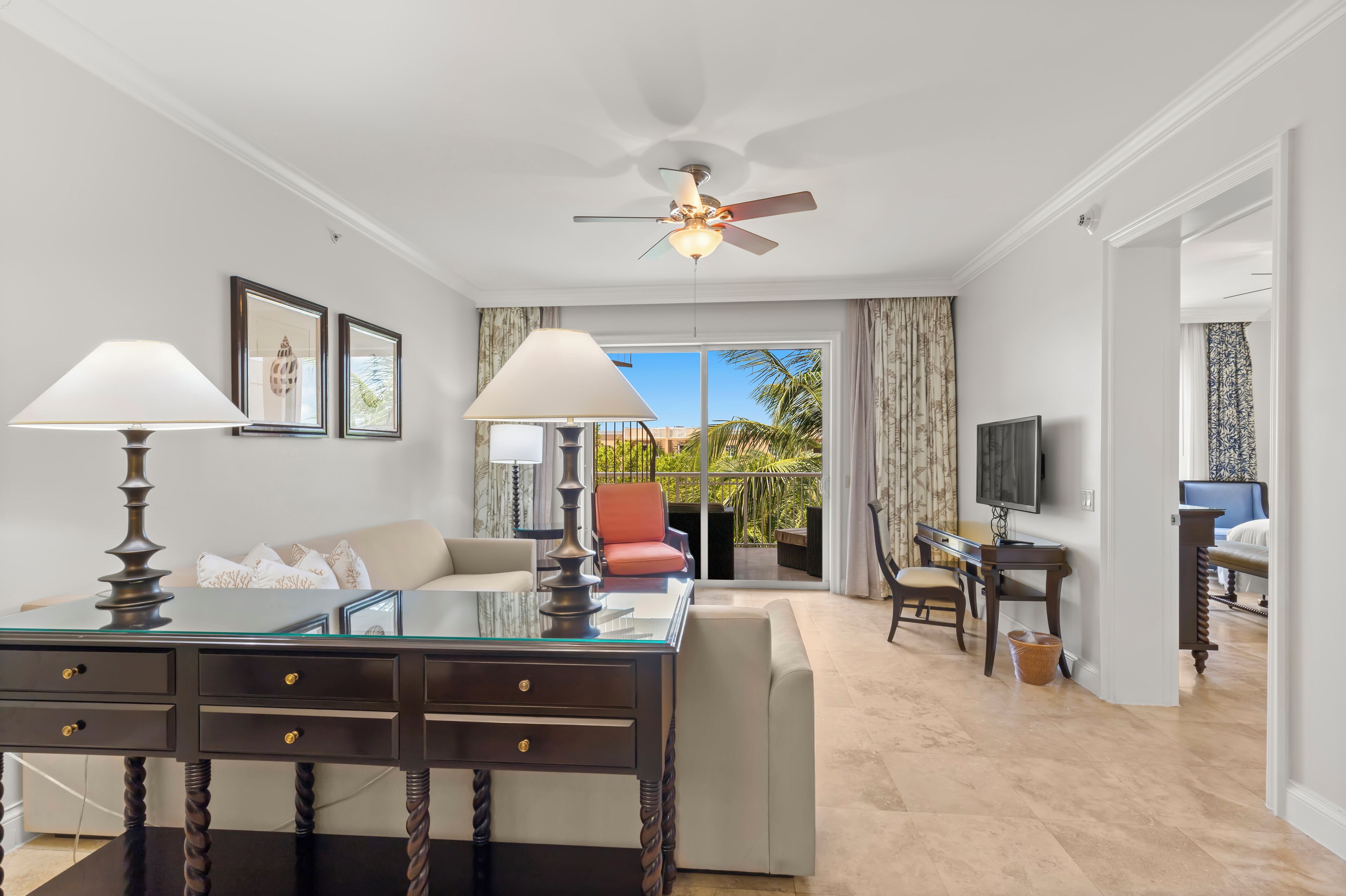 A modern hotel room with a glass desk, two lamps, a sofa, a TV, and a balcony with curtains.