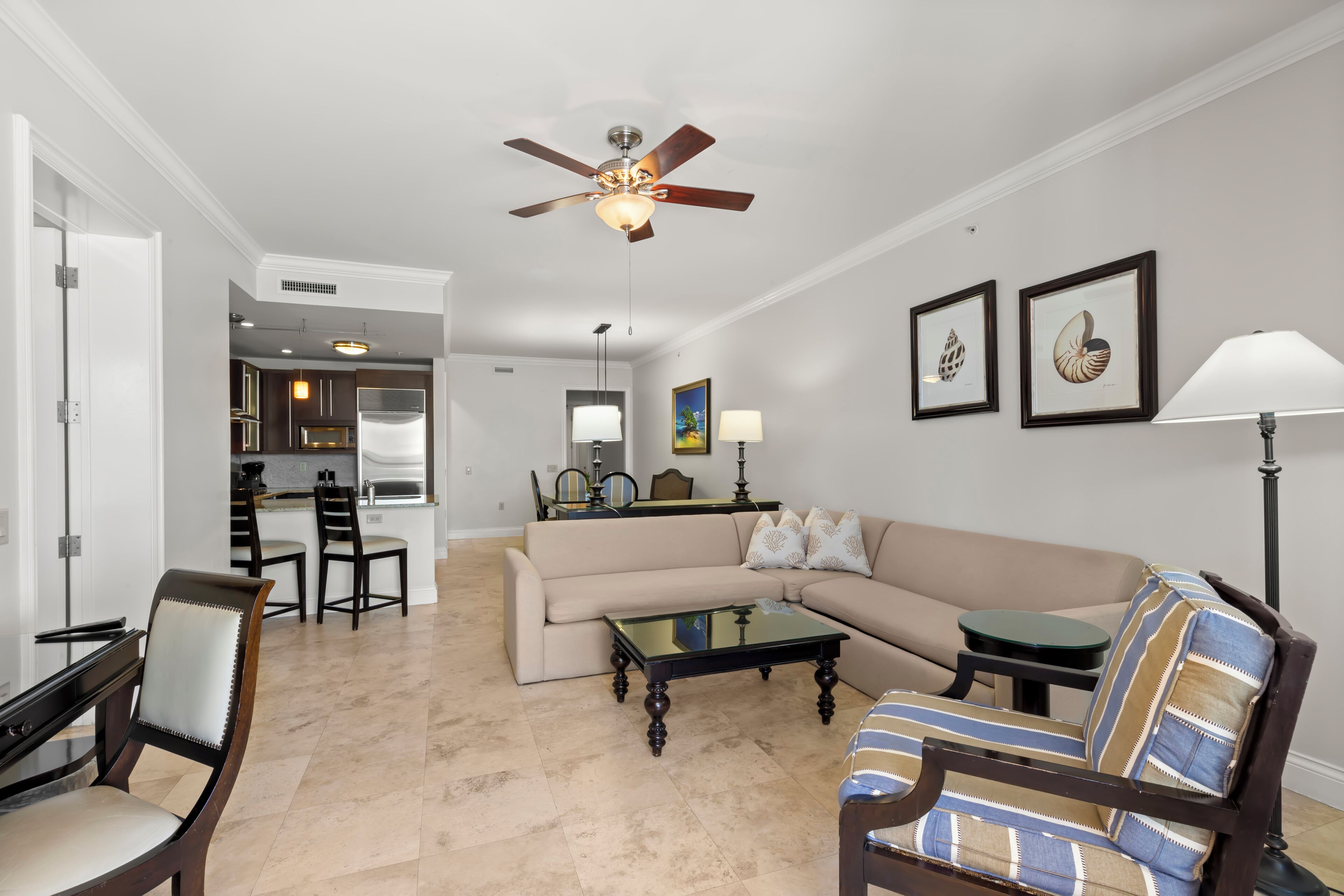 A living room with a beige sofa, a striped armchair, a glass coffee table, and a ceiling fan. There is a kitchen in the background with a dining area.