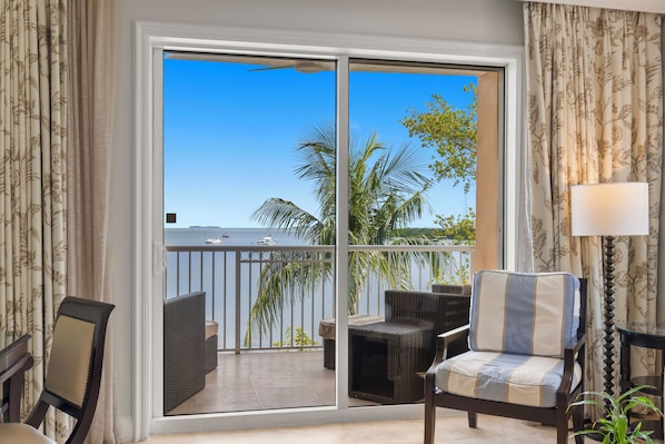 A room with a large sliding glass door leading to a balcony with wicker furniture and a view of palm trees and a body of water.