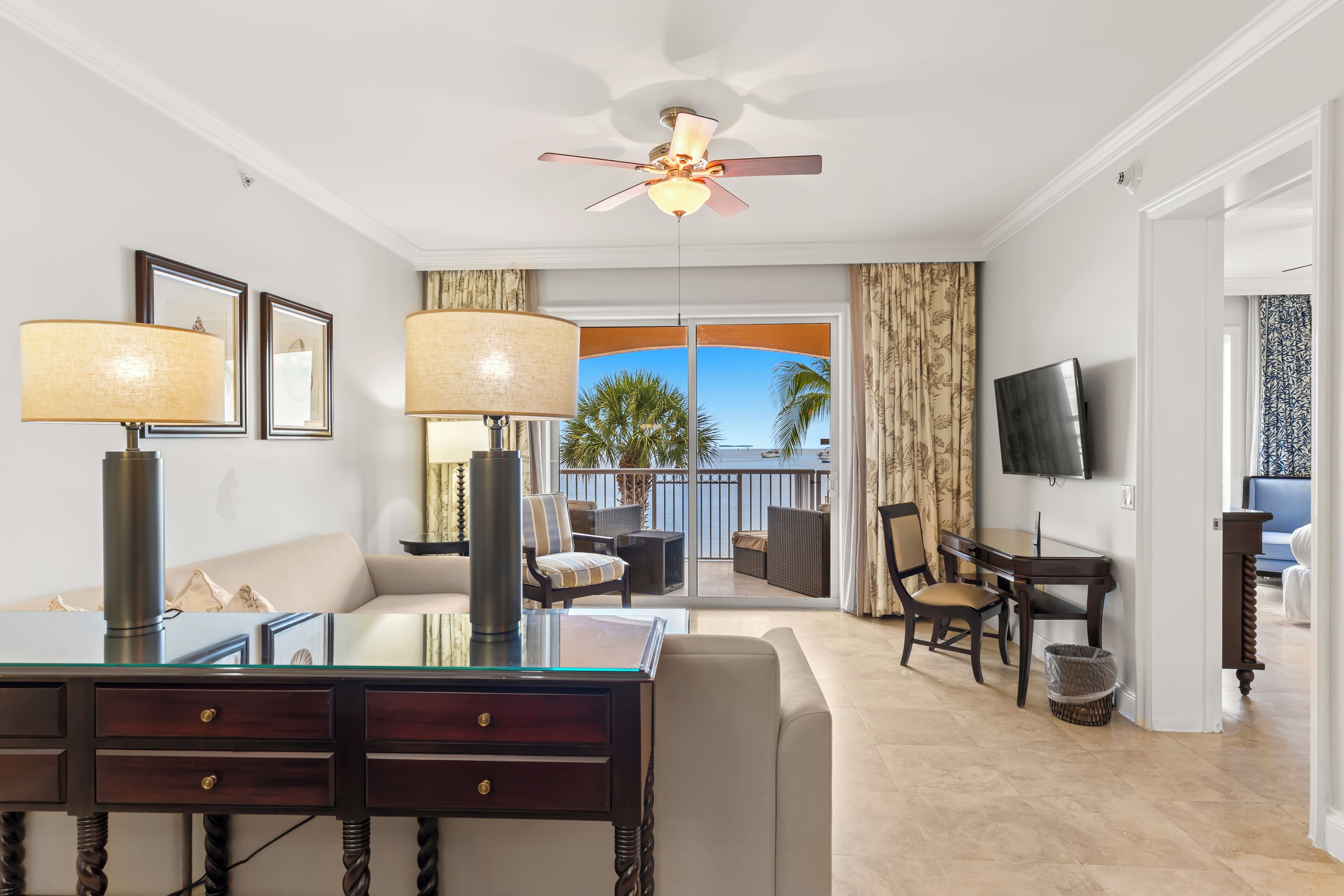 A modern living room with a glass coffee table, dark wooden side tables, a sofa, and a ceiling fan. A balcony with a view of palm trees is visible through the glass doors.