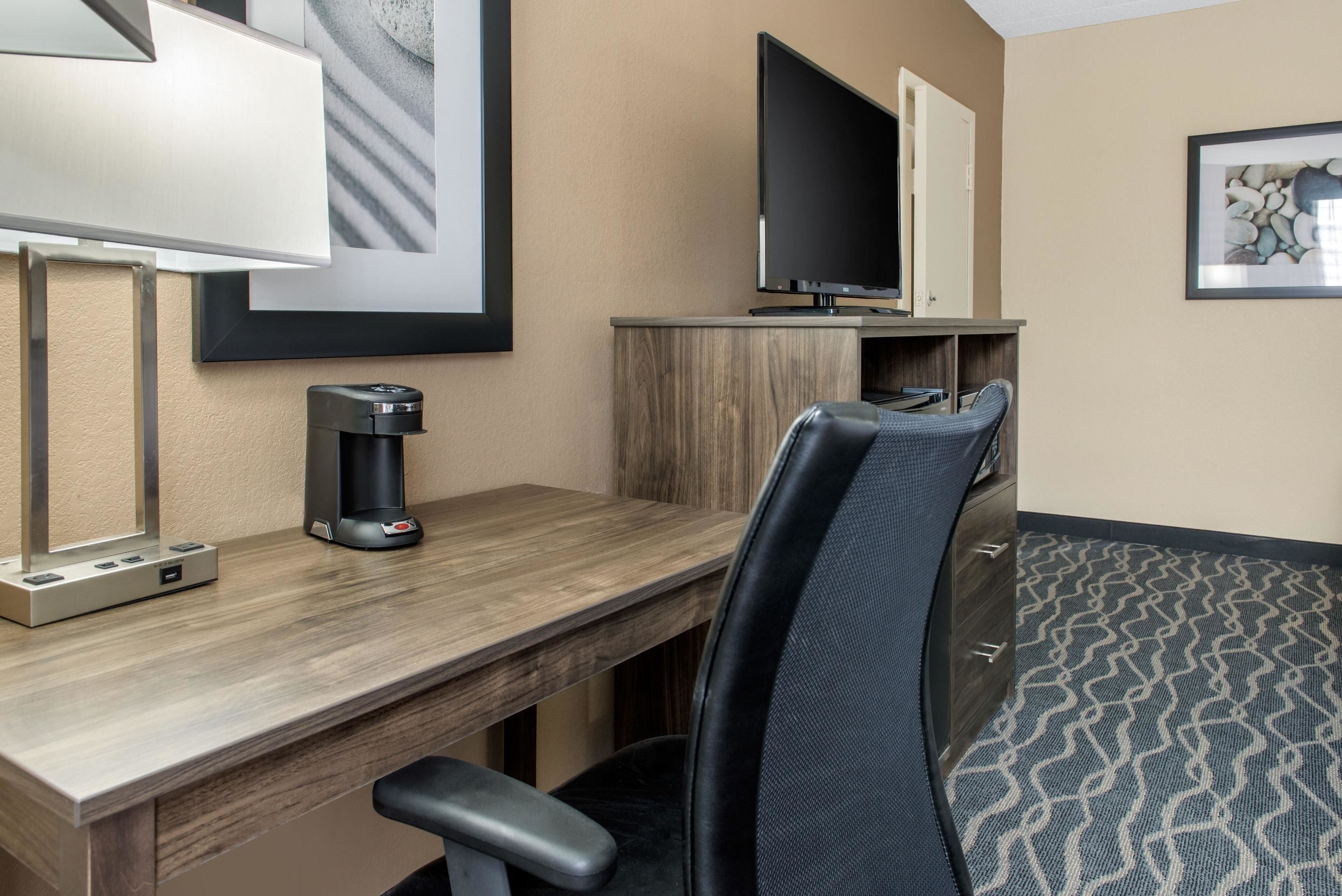 Habitación de hotel con escritorio de madera, una silla de oficina negra, televisor de pantalla plana, cafetera y alfombra estampada.