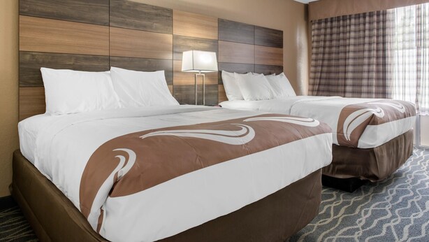 Ein Hotelzimmer mit einem großen Bett, einem kleineren Bett, einem Nachttisch, einer Lampe und einem Fenster mit Vorhängen.