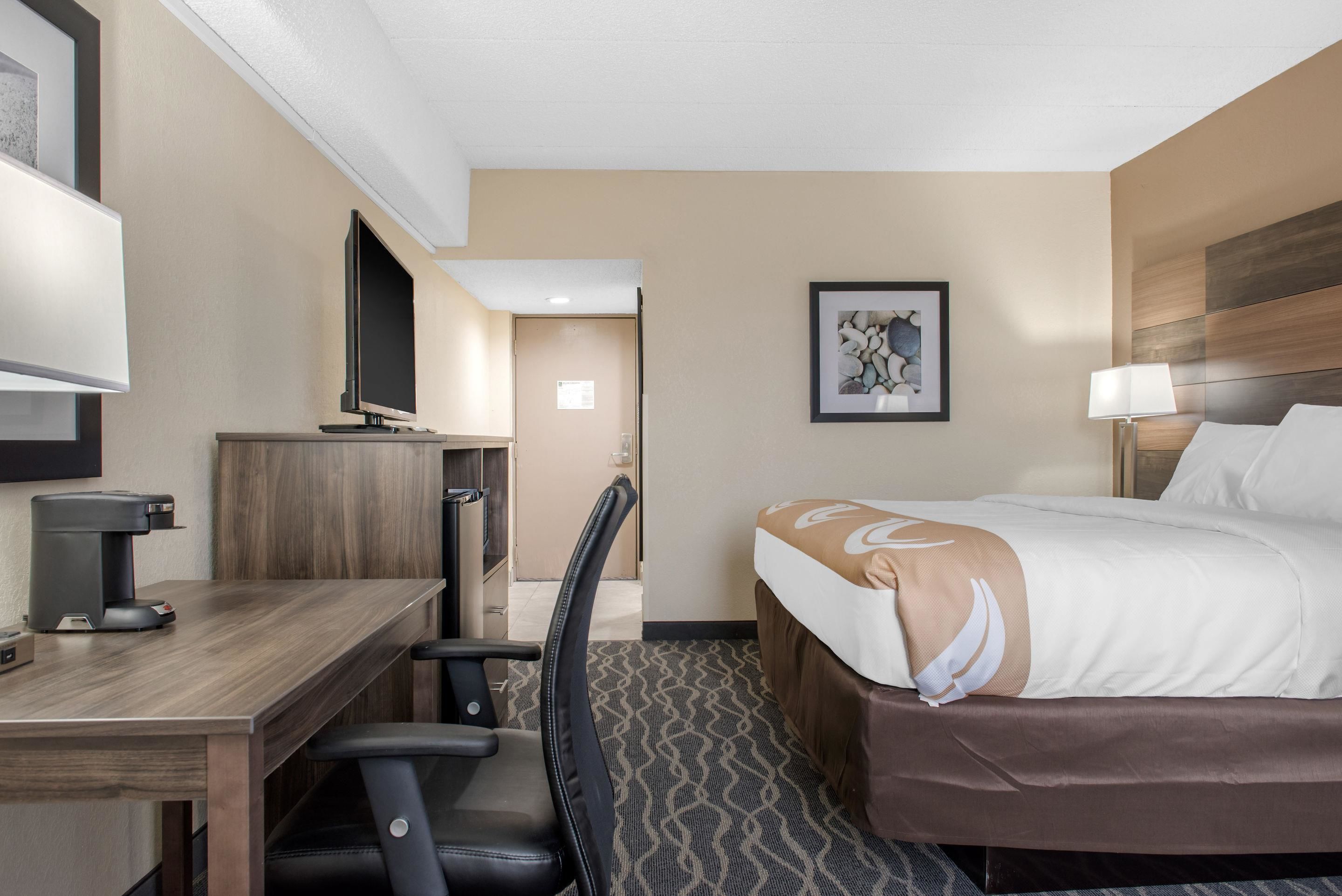 A hotel room with a bed, desk, chair, TV, and artwork on the wall.