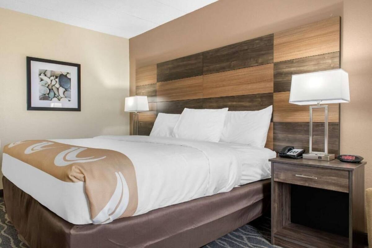 Een hotelkamer met een bed, bedlampjes, een nachtkastje met een telefoon en een wekker, en een lijst aan de muur.
