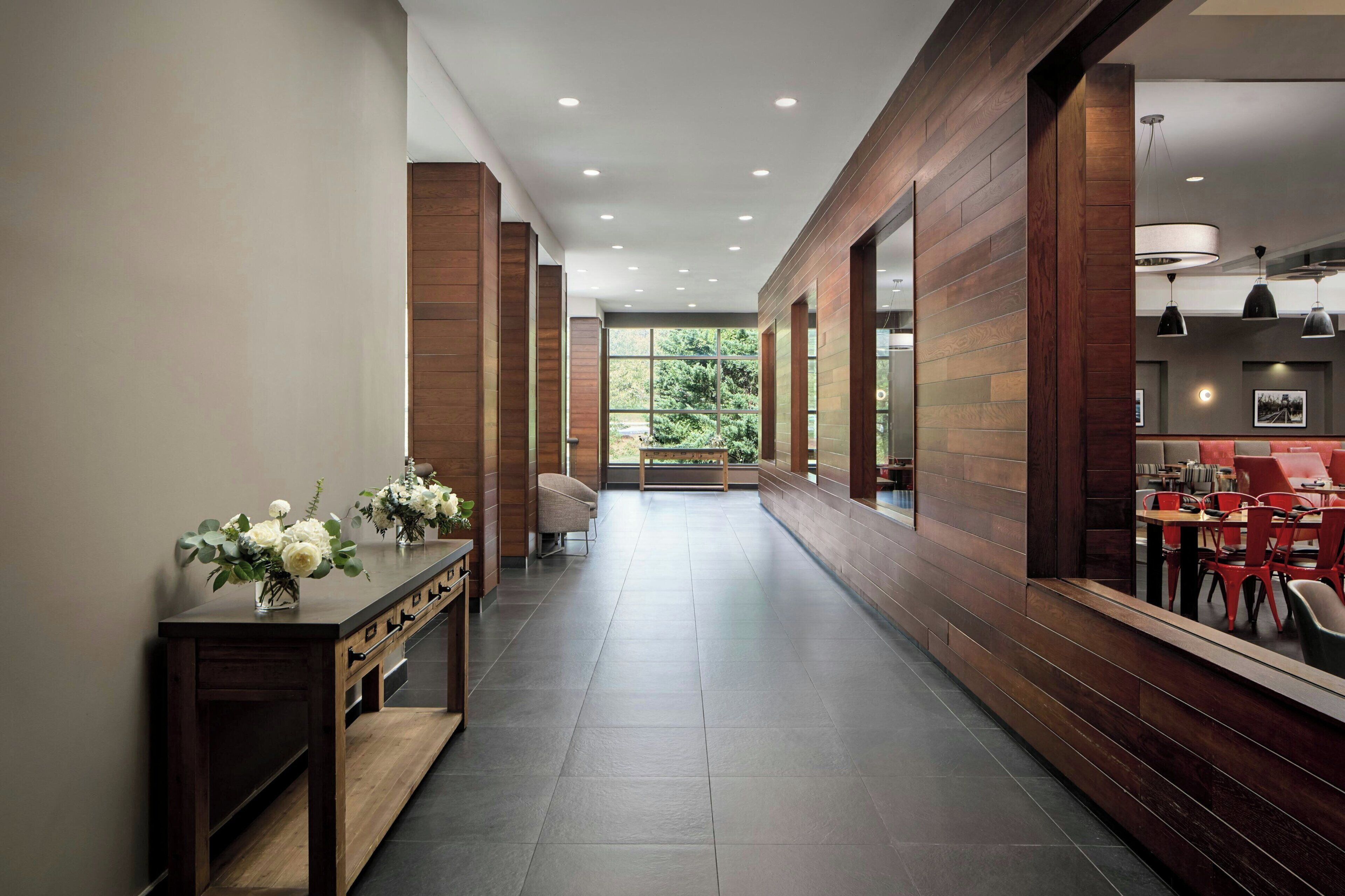 Un pasillo con paneles de madera, un mostrador de recepción con un florero y una zona de estar con mesas y sillas.