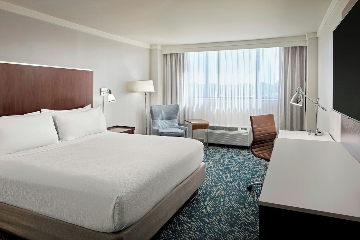 A hotel room with a large bed, a desk, a chair, a lamp, and a view of the city through the window.