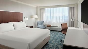 A hotel room with a large bed, a desk, a chair, a lamp, and a view of the city through the window.