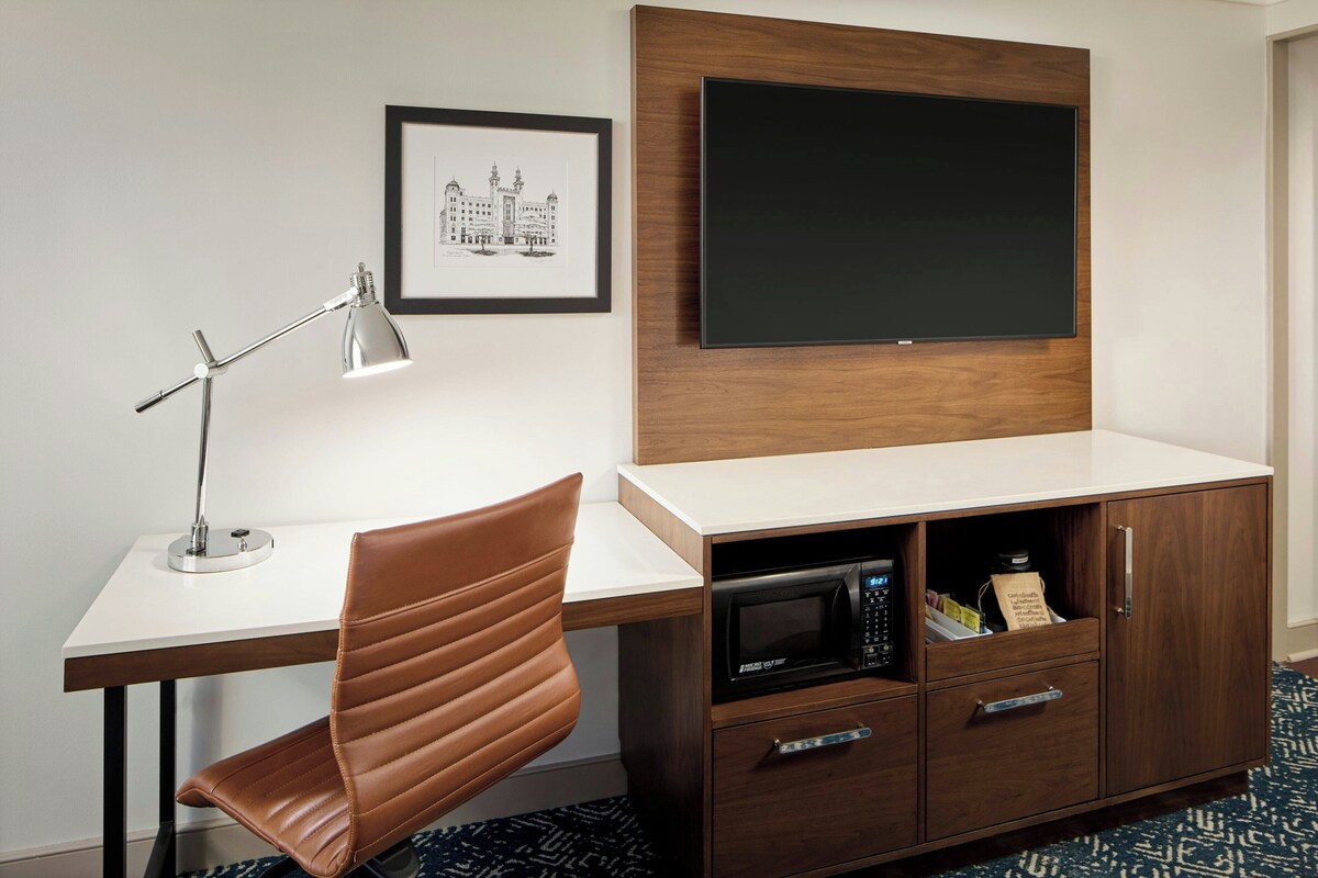 Habitación de hotel con escritorio, una butaca de cuero marrón, un cuadro enmarcado, un televisor de pantalla plana y un microondas.