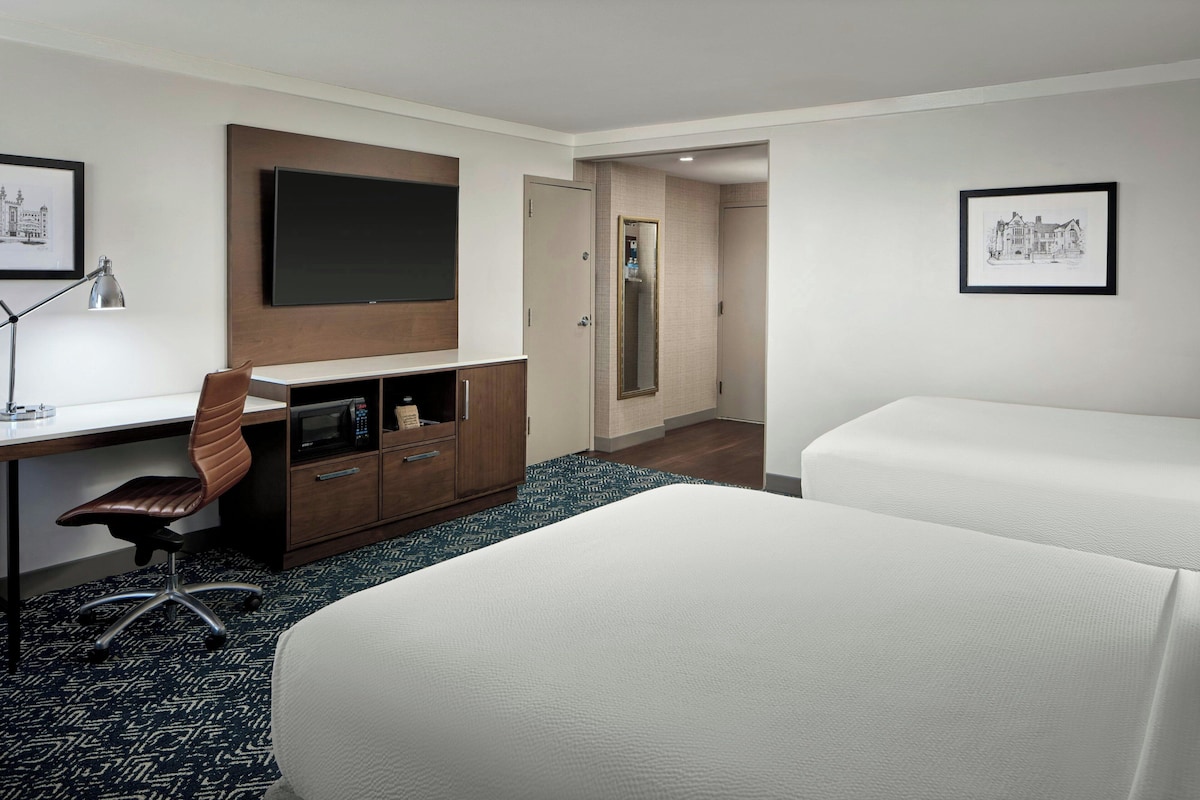 Habitación de hotel con escritorio, silla, dos camas, espejo y cuadros en las paredes.