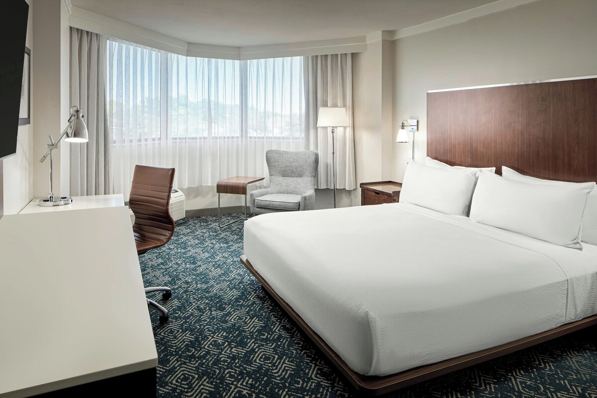 A hotel room with a large bed, a desk, a chair, a lamp, and a view of the outdoors through the window.