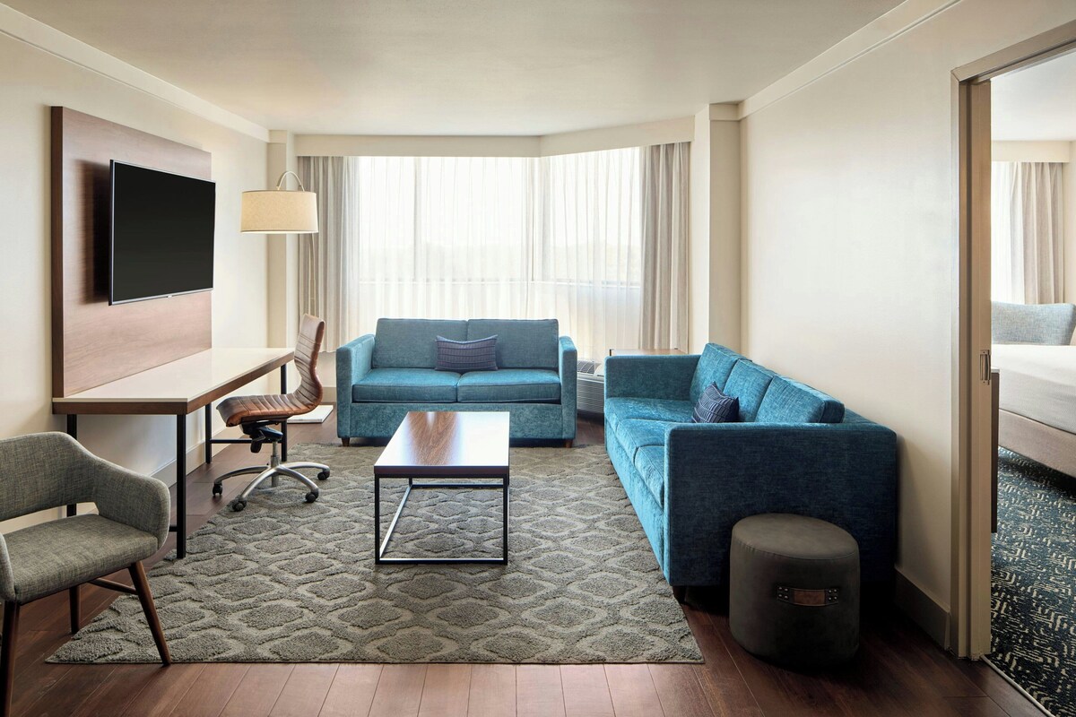 A hotel room with a flat-screen TV, a desk with a chair, two sofas, a coffee table, and a small ottoman.