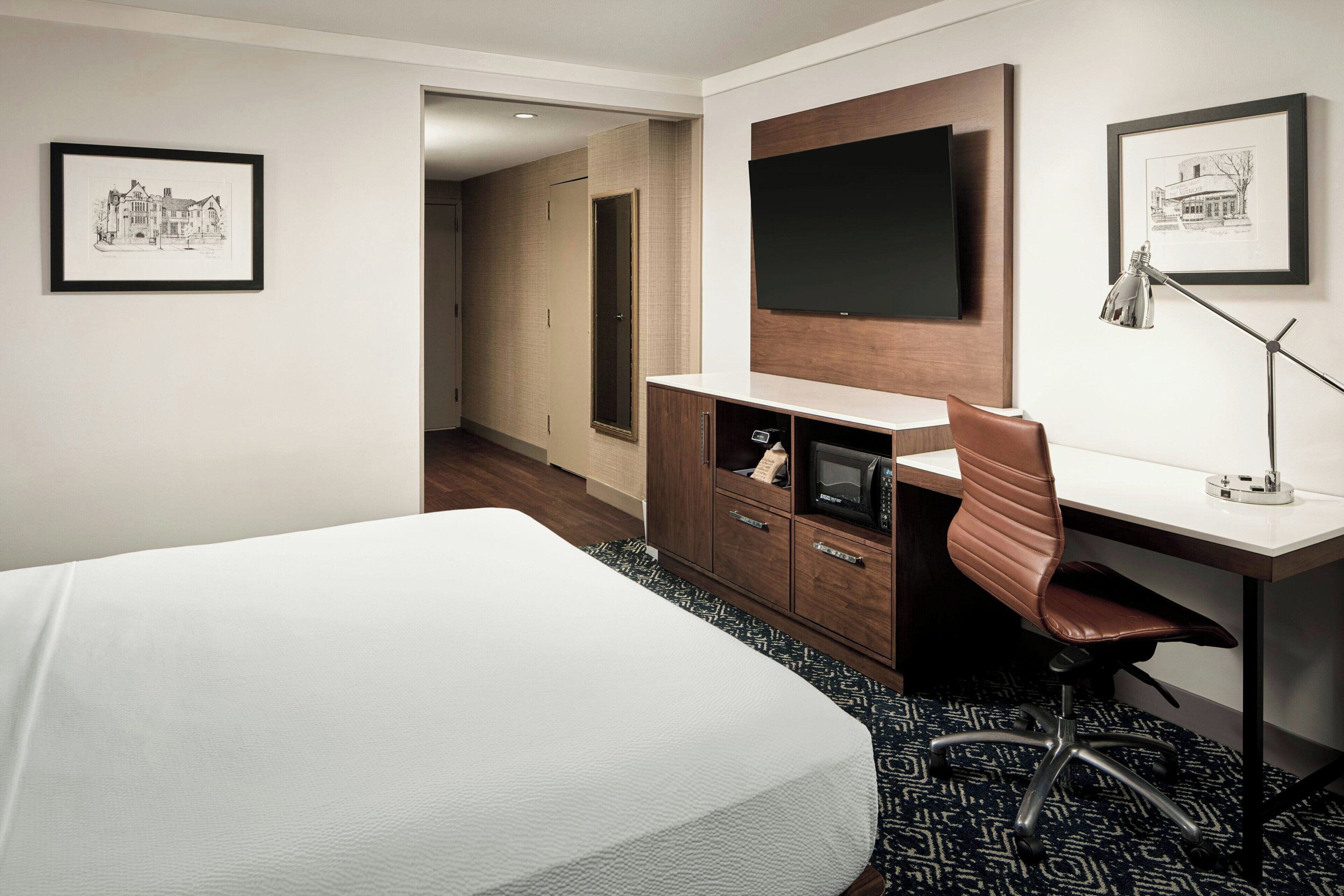 Ein Hotelzimmer mit einem Bett, einem Schreibtisch, einem Sessel, einem Fernseher und einem gerahmten Bild an der Wand.