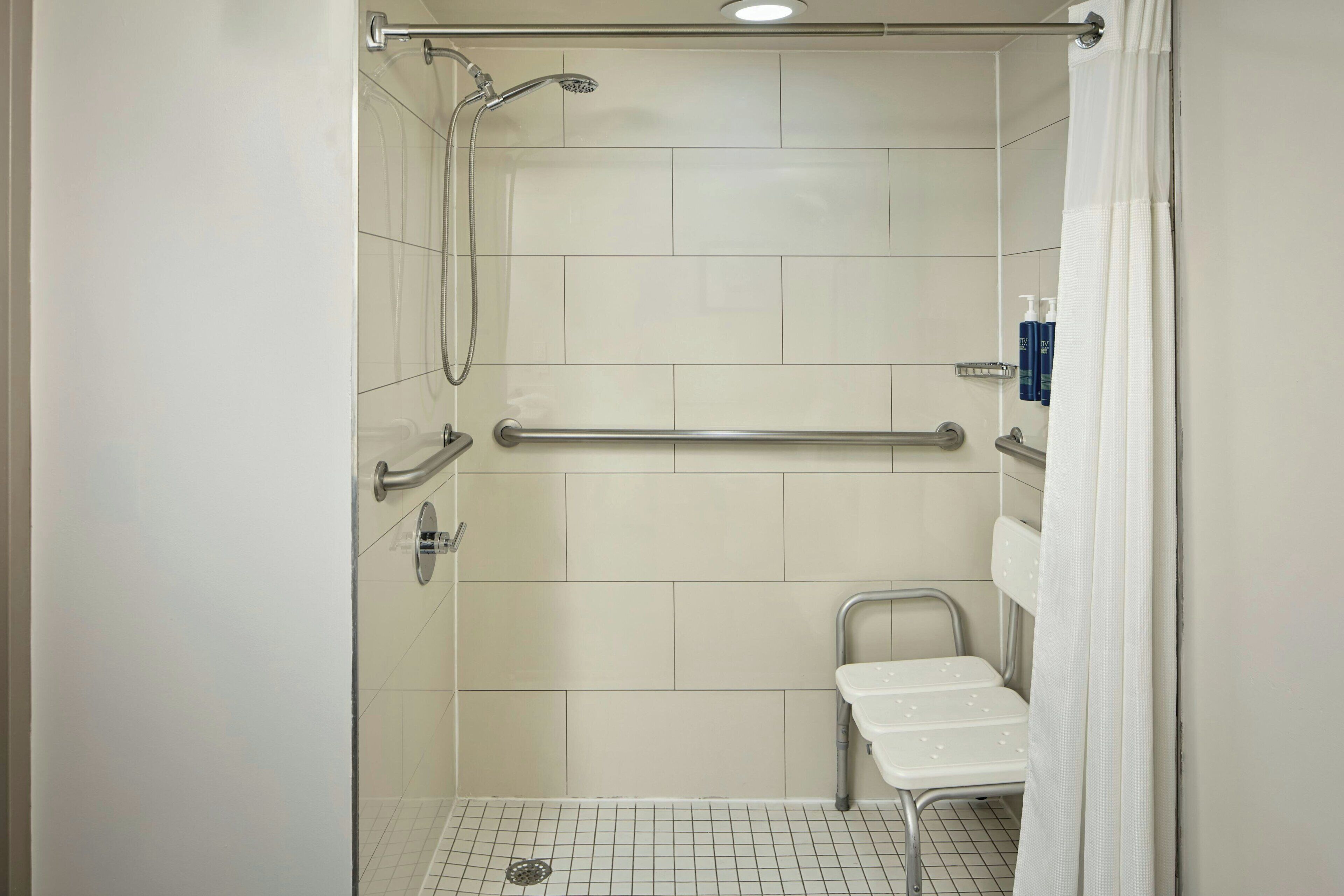 Zona de ducha con barra de apoyo, ducha de mano y un inodoro con asiento de seguridad.