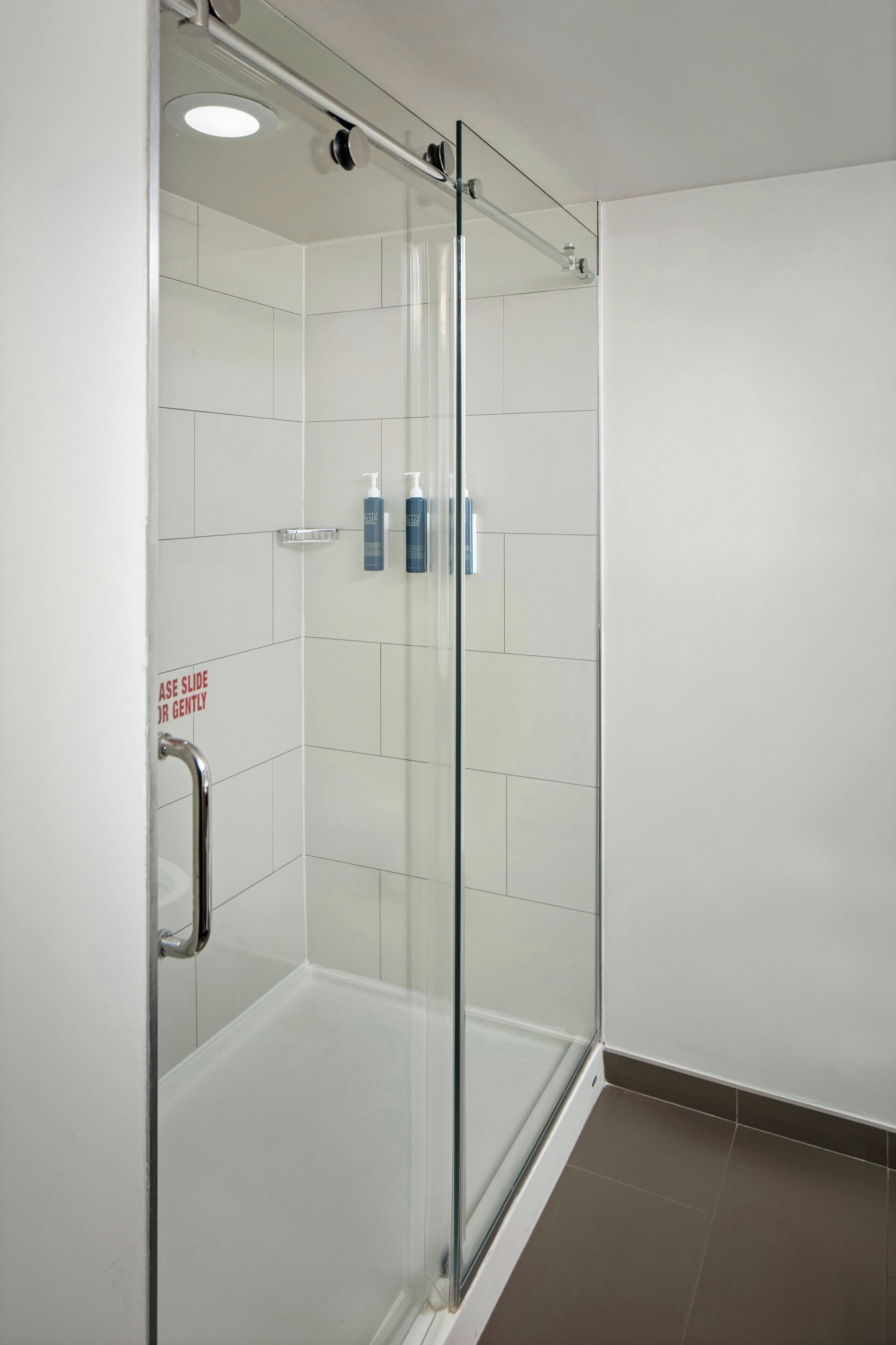 A glass-enclosed shower stall with a handrail, a tiled floor, and a sign advising to 'ASE SLIDE DR GENTLY'.