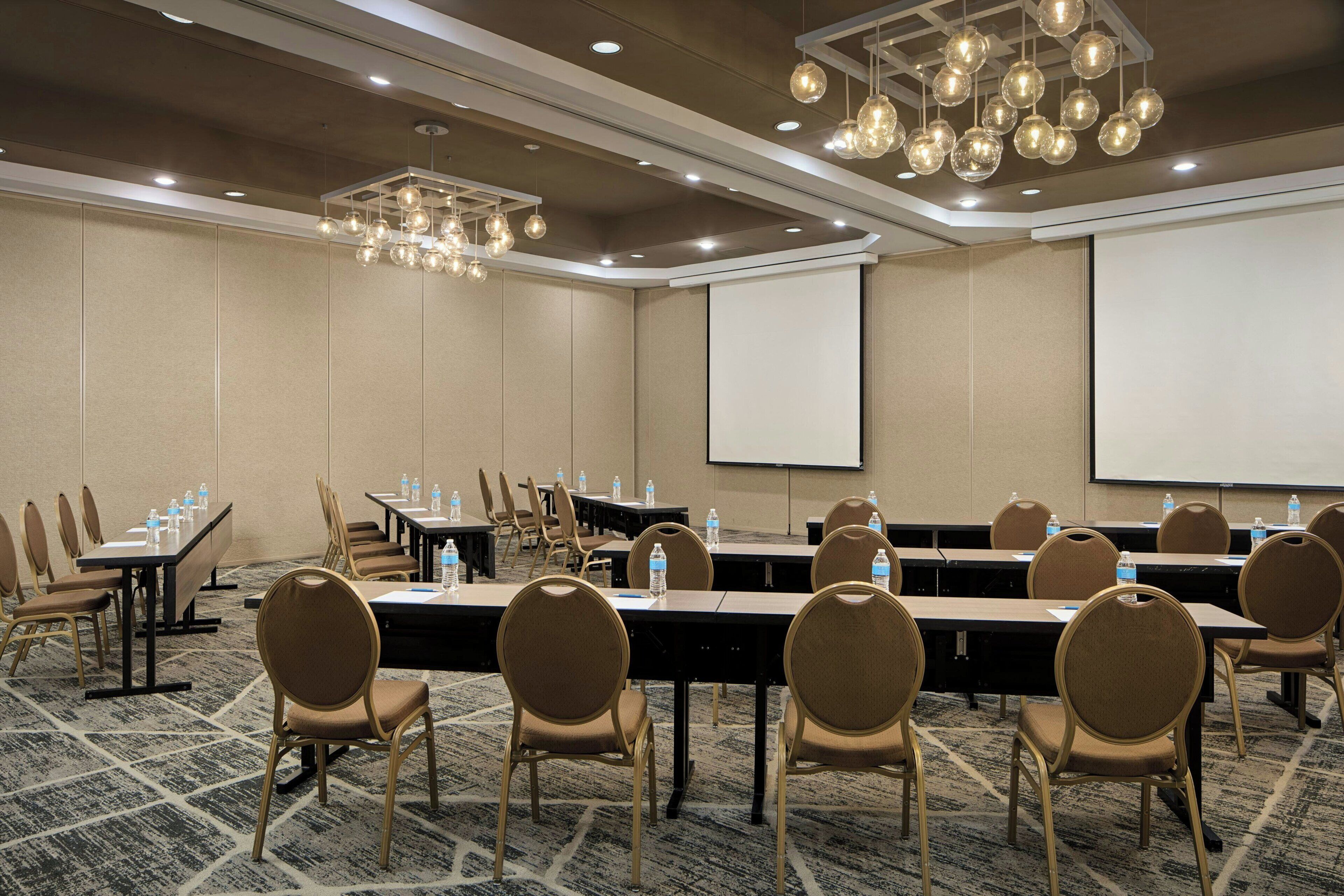 Un salón de conferencias con largas mesas, sillas, botellas de agua, una pantalla de proyector y un moderno candelabro.