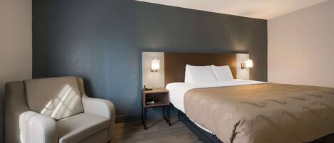 Quarto de hotel com cama, uma cadeira, mesa de cabeceira e duas luminárias fixadas na parede.