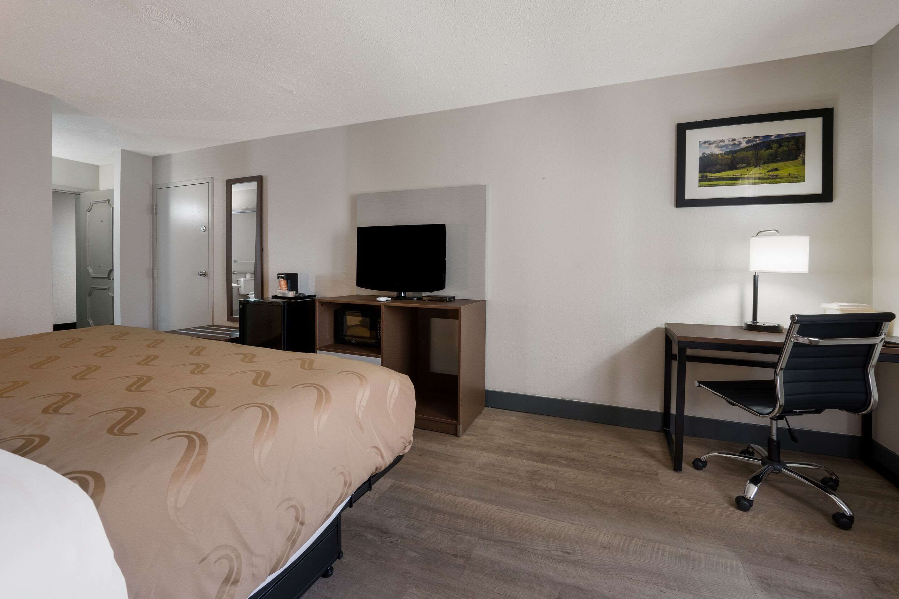 A hotel room with a bed, a desk with a chair, a television, and a framed picture on the wall.