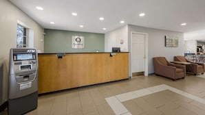 A reception area with a wooden reception desk, an ATM, and a sign that says "Quality by Choice Hotels." There are wicker chairs and a door in the background. - Quality Inn University Area (Farmville)