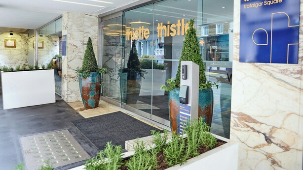 A modern hotel lobby with a reception desk, glass doors, and potted plants. - Thistle Trafalgar - Leicester Square (London)