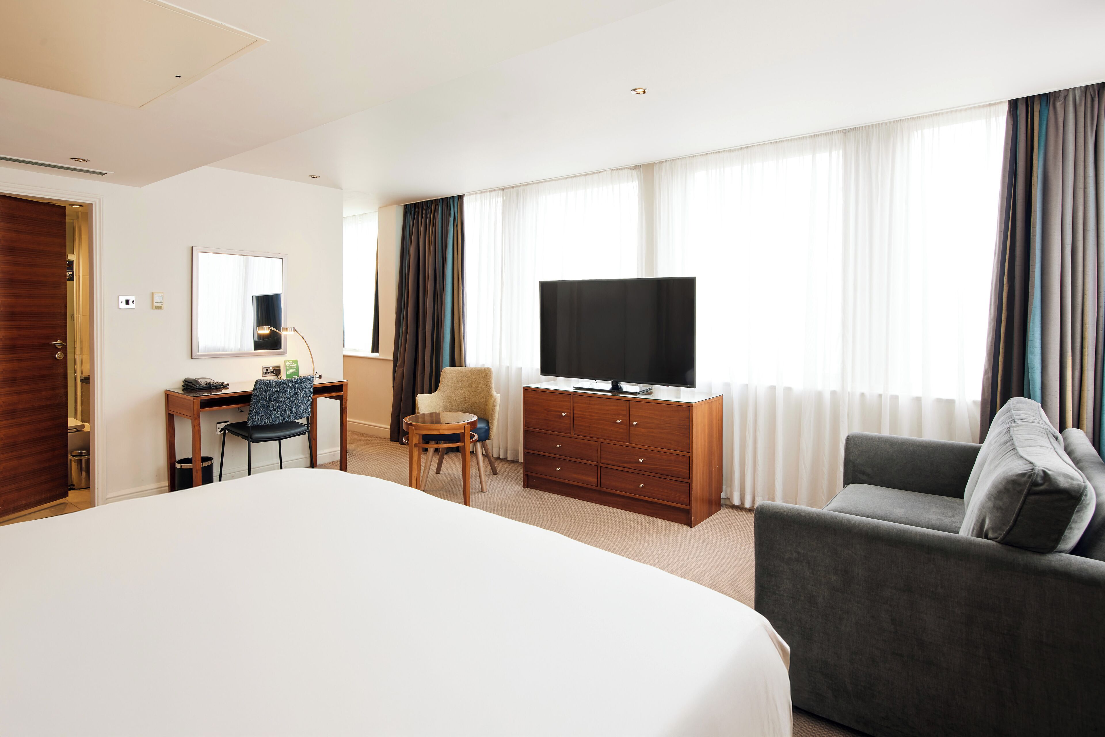 A hotel room with a large bed, a TV on a wooden cabinet, a desk with a chair, a sofa, and a window with curtains.
