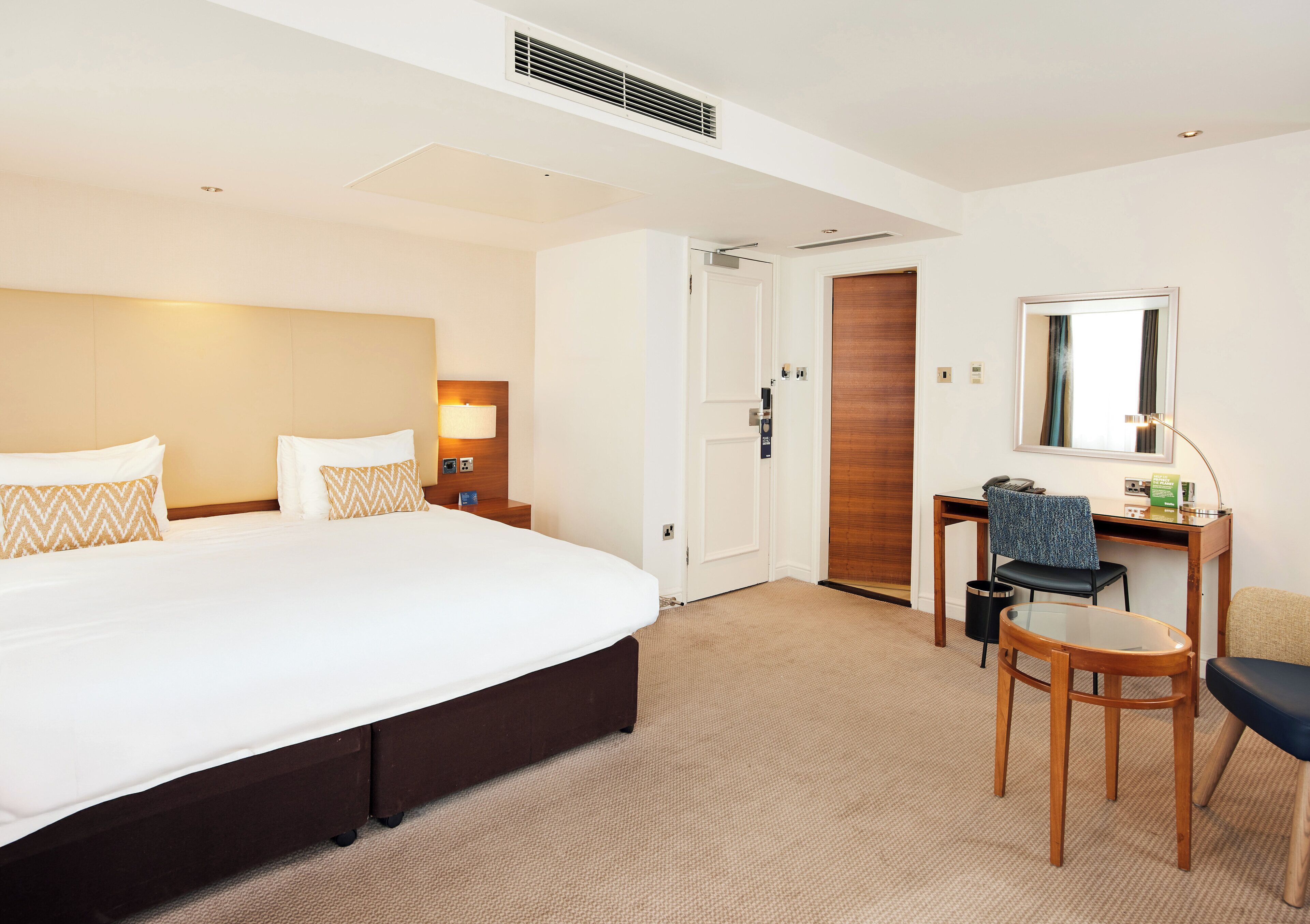 Hotelkamer met een groot bed, een bureau met stoel, een spiegel en een deur die naar een andere kamer leidt.
