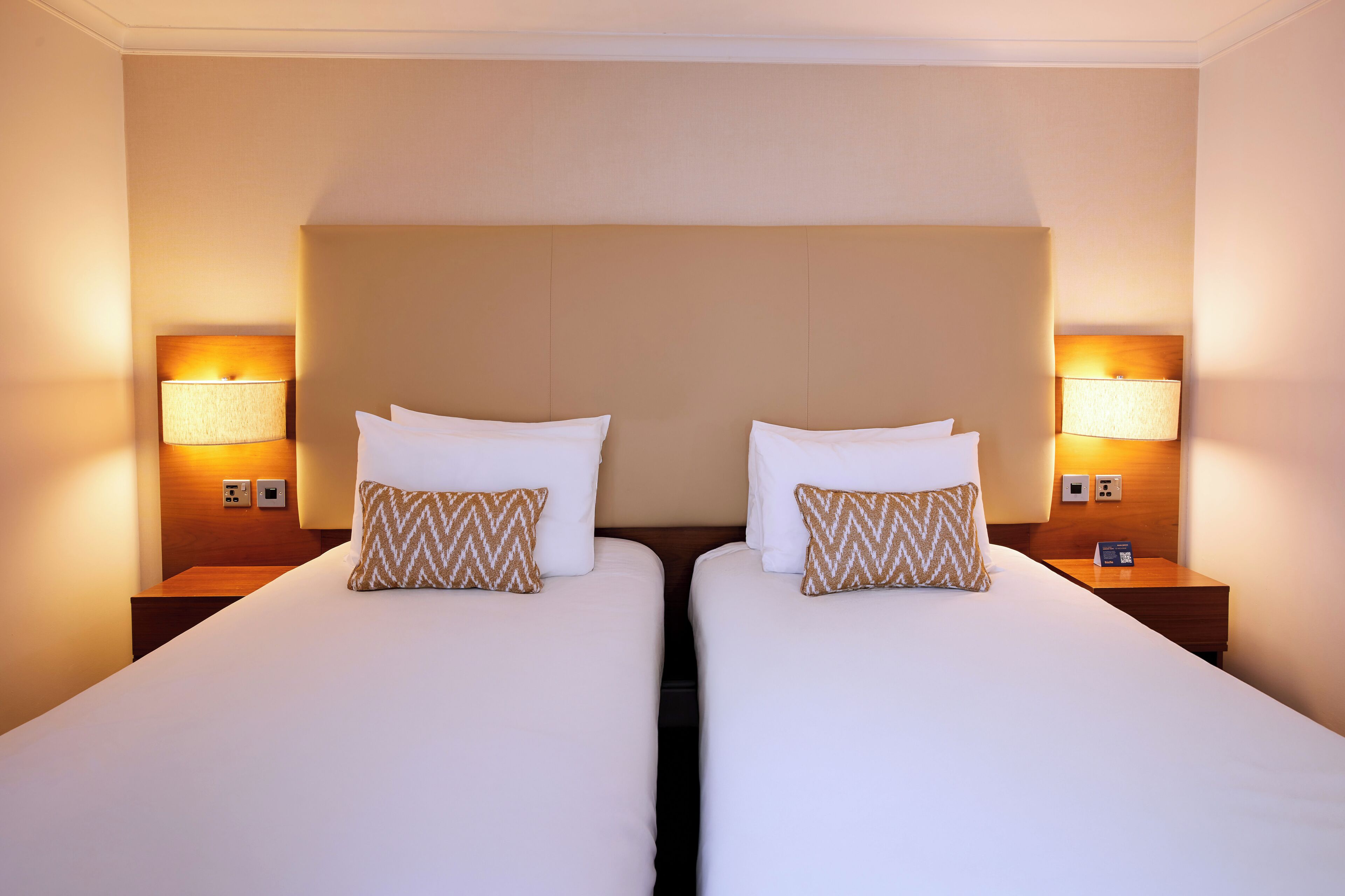 A hotel room with two single beds, each with a wooden nightstand, a lamp, and a patterned pillow.
