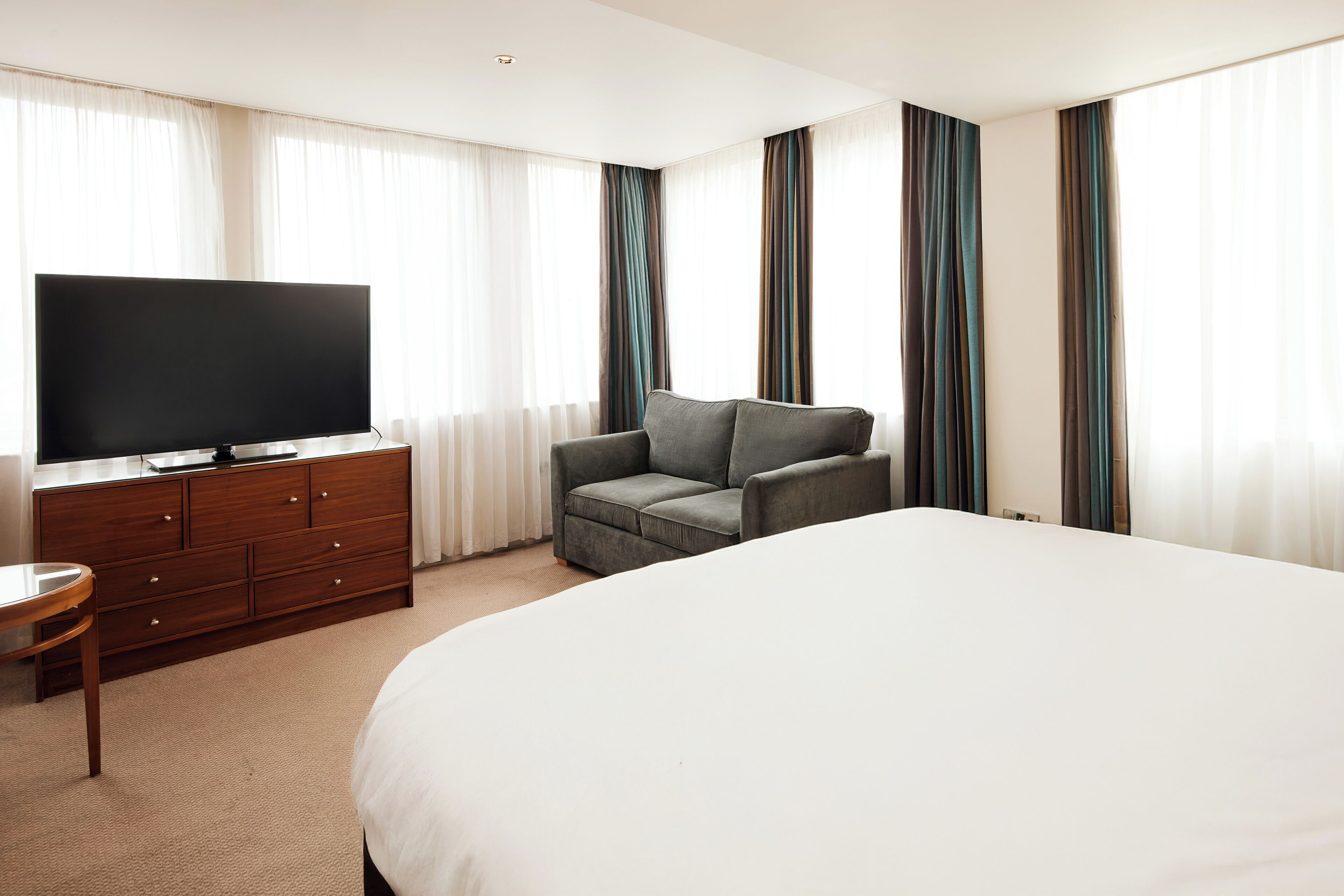 A hotel room with a large bed, a TV on a wooden dresser, a gray armchair, and curtains on the windows.