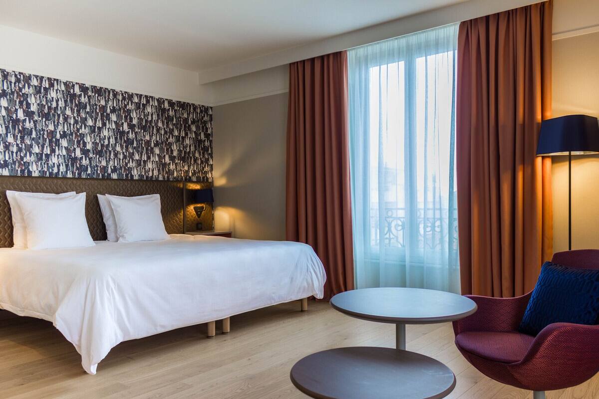 Een hotelkamer met een bed, een ronde tafel, een stoel en een groot raam met gordijnen.