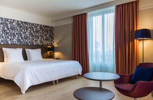 Habitación de hotel con cama, mesa redonda, una silla y un ventanal con cortinas.