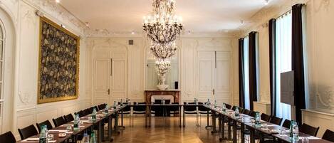 A formal meeting room with a long table, chairs, a chandelier, and a large painting on the wall.