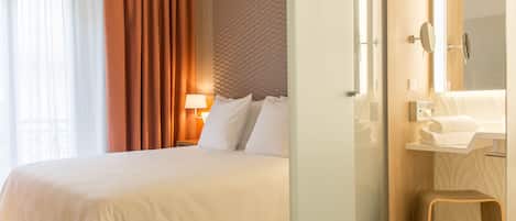 Una habitación de hotel con una cama grande, una lámpara de noche, un espejo y un taburete pequeño.